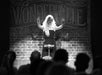 A performer with long hair stands on stage in front of a "Wonderville" sign, hands raised, in front of an audience.
