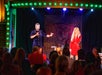 Two performers on stage entertain an audience. One is dressed in a blue suit with a bandaged head; the other wears a red dress. The audience watches under colorful stage lights.