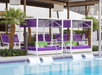 Poolside scene with purple lounge chairs and cabanas featuring green cushions. Palm trees are visible, and partially submerged lounge chairs are in the pool.