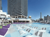 Hotel pool area with submerged lounge chairs, purple daybeds, and large building in background. Parisian-style tower visible in the skyline.
