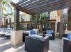 Outdoor patio with wicker furniture, cushions, and a wooden pergola. There are palm trees in the background and a two-story building.