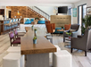 Spacious modern lounge with wooden tables, brown and gray chairs, blue cushioned seating, plants, and a staircase in the background. Bright lighting and neutral tones enhance the decor.