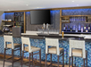 A modern bar with four high chairs, a flat-screen TV, a chalkboard menu, and shelves with bottles and glasses. The bar has blue mosaic tiles and various beverage dispensers.