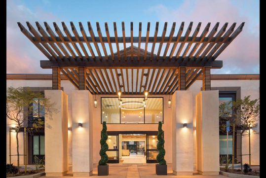 Modern building entrance with a wooden pergola, large glass doors, and two symmetrical topiary plants. The facade is white with black trim, set against a pink and blue sky.