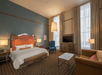 A hotel room with floor to ceiling windows, a white king bed, blue accent chair, brown love seat, coffee table, and a dresser with a TV on it.