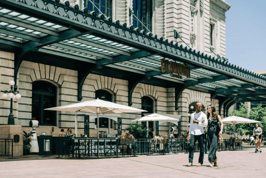 A covered patio with an iron fence around it and large white umbrellas over the tables on a sunny day with people walking by at the The Crawford Hotel.