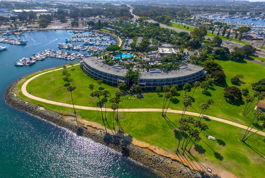 Aerial view of a curved waterfront building with a pool, surrounded by lush greenery and palm trees, adjacent to a marina filled with boats. Paths wind along the shoreline.