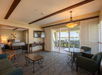 Hotel room with a seating area, bed, wall-mounted TV, and sliding glass doors leading to a balcony with a water view.