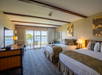 Hotel room with two beds, a flat-screen TV, dresser, decorative artwork, and a sliding glass door leading to a balcony with chairs and a lake view.