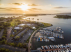 Aerial view of a marina at sunset with boats docked, surrounded by green spaces, palm trees, and water.