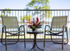 Two chairs face a small table with a colorful flower arrangement and a white cup on a balcony with a railing, overlooking greenery and distant water.