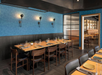 Restaurant interior with wooden tables and chairs, set for dining. Blue textured walls with three wall lights. Open view to booths and shelving in the background.
