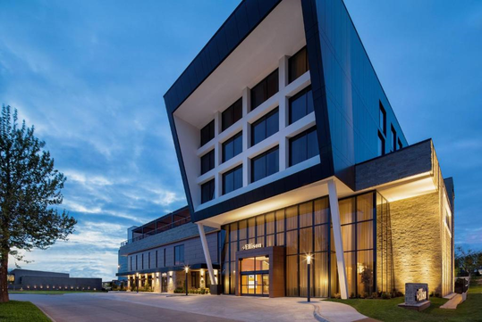 Modern hotel building with angular design, large windows, and a well-lit entrance at dusk. A sign with "Hilton" is visible near the entrance.
