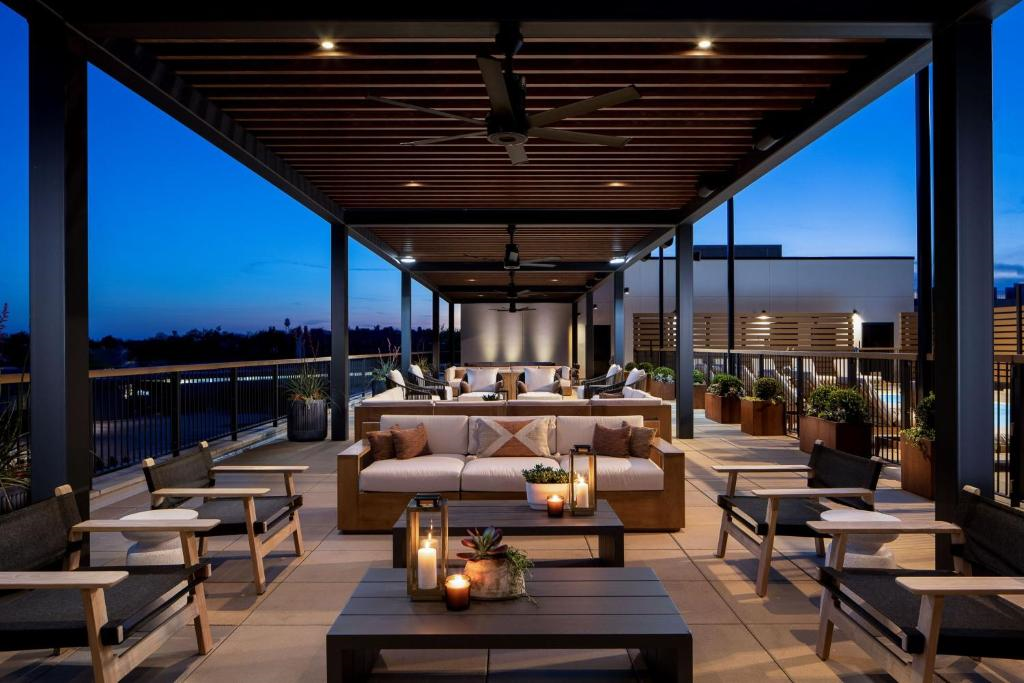 Rooftop lounge with modern sofas, cushioned seating, and tables under a wooden pergola at dusk. Candles and potted plants are placed around. Urban skyline visible in the background.