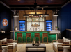 Modern bar with green-tiled counter, wooden stools, liquor shelves, and ambient lighting. Two white sofas face the bar, creating a cozy seating area.