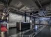 A modern lobby features a sleek black counter, geometric floor patterns, and metallic ceiling with overhead pipes. Wall art includes a diagram and a collection of small lights.