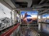A modern exhibition space featuring a large photograph of a hand holding a lens with a cityscape inside, surrounded by aviation-themed wall art.