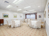 A banquet room with round tables covered in white tablecloths and surrounded by beige chairs. Framed paintings hang on the walls.