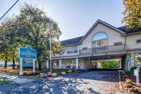 Street view of The Farmington Inn, featuring a two-story building with a parking area beneath. An American flag is displayed, and landscaped flower beds frame the entrance.
