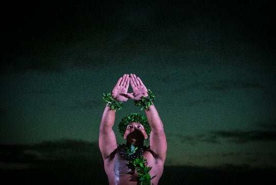 A male hula dancer with his hands together toward the sky with clouds in the background at The Feast at Mokapu Luau in Wailea, Hawaii, USA.