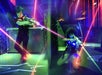 Two kids wearing laser tag vests play in a neon-lit arena, aiming laser guns as colorful beams and glowing lights fill the scene.