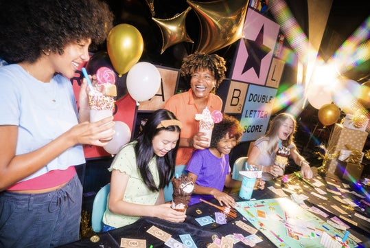 Five people sit at a table playing a board game and holding milkshakes, surrounded by balloons and colorful decorations.