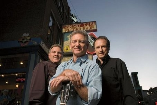 Three men stand in front of Ernest Tubb Record Shop, one holding a guitar, with a cityscape in the background.