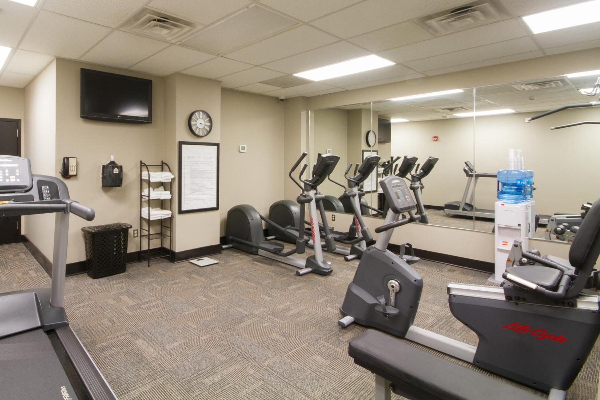 Small gym room with exercise bikes, treadmill, a wall mirror, TV, clock, water dispenser, and shelves with towels against beige walls and carpeted floor.