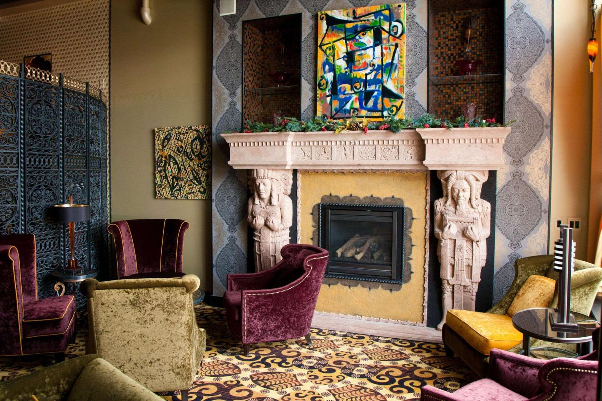 A lounge with colorful armchairs, a patterned carpet, ornate decor, and a fireplace with two carved figures and abstract art above the mantel.
