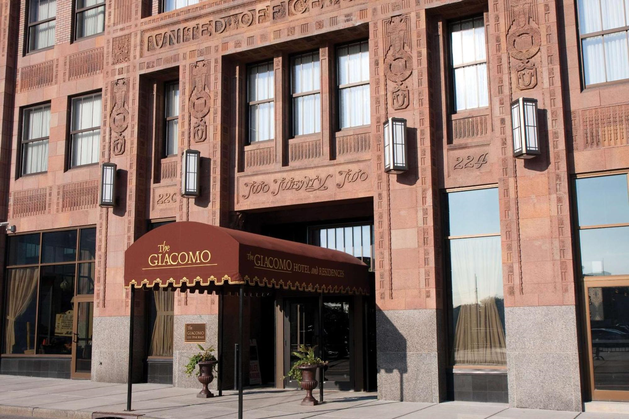 Entrance of The Giacomo hotel with a brown canopy, large windows, and detailed brickwork on the building facade.