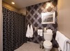 A bathroom with patterned wallpaper, a shower with a black and gray curtain, white towels and robes, a toilet, and framed wall art above the toilet.