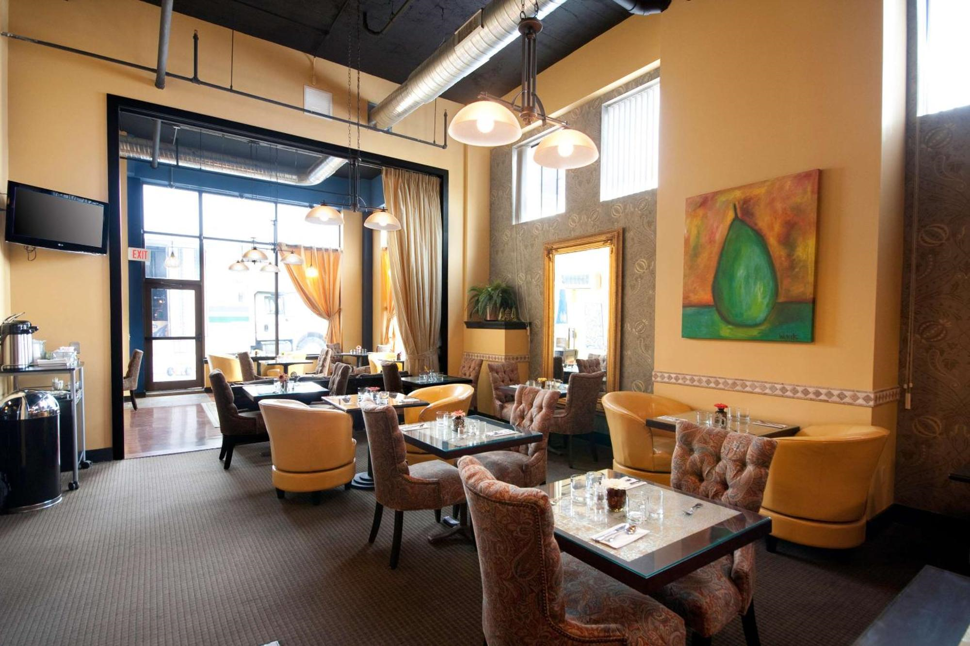 A cozy restaurant dining area with yellow walls, upholstered chairs, glass tables, wall art, large windows, and modern pendant lighting.