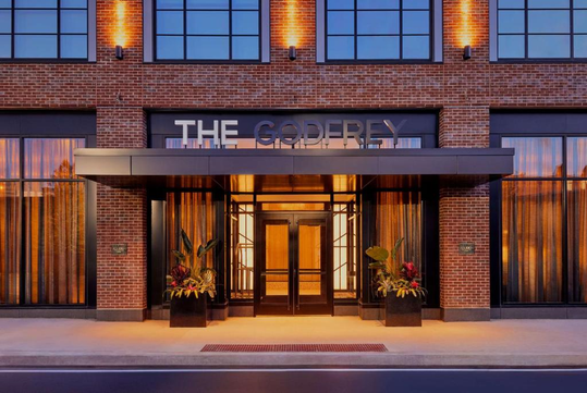 Brick building entrance with glass doors and "The Godfrey" sign above. Large windows flank the entrance, adorned with potted plants. Warm lighting illuminates the exterior.