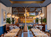 Stylish restaurant interior with modern lighting, wooden tables set with glassware, and green plants. Booth seating lines the walls, creating a warm and inviting atmosphere.