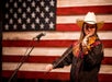 A person in a cowboy hat plays a violin next to a microphone, with an American flag in the background.