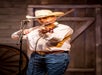A man in a cowboy hat and jeans plays the violin on stage, standing in front of a microphone. A wooden door and wagon wheel are visible in the background.
