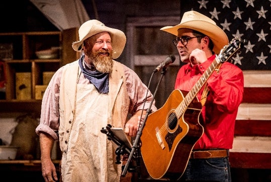 Two men in cowboy hats, one with a guitar, perform on stage. An American flag is in the background.