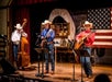 Three musicians in cowboy attire perform on stage with guitars and a double bass, with an American flag and wagon wheel in the background.