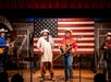 Four people wearing cowboy hats perform on stage with an American flag backdrop and musical instruments.