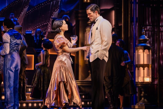 A woman in a gold dress talks to a man in a white suit at a formal event. People are socializing in the background, with an orchestra playing on stage.