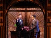 Two men in suits engage in conversation on a stage with an art deco backdrop, one seated on a desk and the other standing, gesturing with one hand.