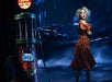 A woman in a patterned orange dress stands near a vintage gas pump with an "OPEN GAS" sign, against a dark, dramatic backdrop.
