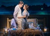 Two actors in period costumes kneel closely facing each other on a bed, with soft lighting and a nighttime background scene behind them.