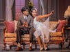 Two actors in vintage attire sit on a patterned sofa on stage; the woman gestures outward while the man looks at her, with a decorative set and window in the background.