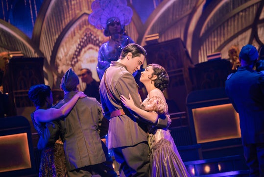 A man in a military uniform and a woman in a gown dance closely on stage, surrounded by other dancing couples and elaborate Art Deco-style set pieces.