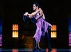 Two dancers perform a dramatic pose on stage, with the man supporting the woman as she leans back. The background features illuminated architectural elements.