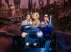 Three people enjoying a ride in a vintage car, set against a theatrical backdrop of a cityscape.