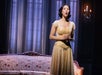 A woman in a formal gown sings on stage in front of a decorative couch and tall windows with curtains. A chandelier is visible in the background.