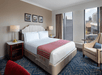 Hotel room with a large bed, red throw, two bedside tables with lamps, an armchair, and art on the wall. Large windows offer a city view.