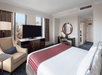 Hotel room with a bed, armchair, TV, and desk. Large windows offer a city view, and a round mirror hangs above the desk. The decor is modern and neutral-toned.